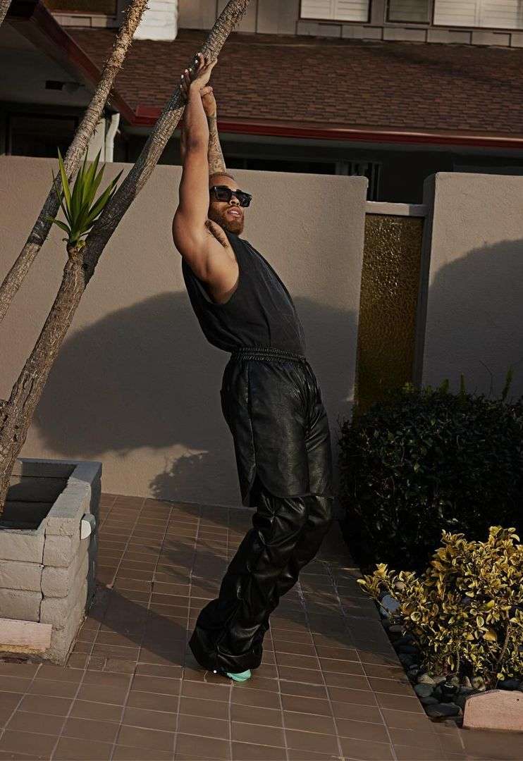 Stylish athletic man in black tank top and pants posing outdoors, near a tree and building with a stone walkway, and wearing sunglasses.
