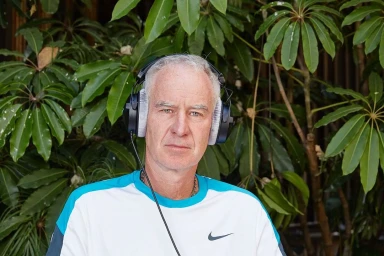 Middle-aged man wearing black over-ear headphones and a white and blue athletic shirt against lush tropical foliage.