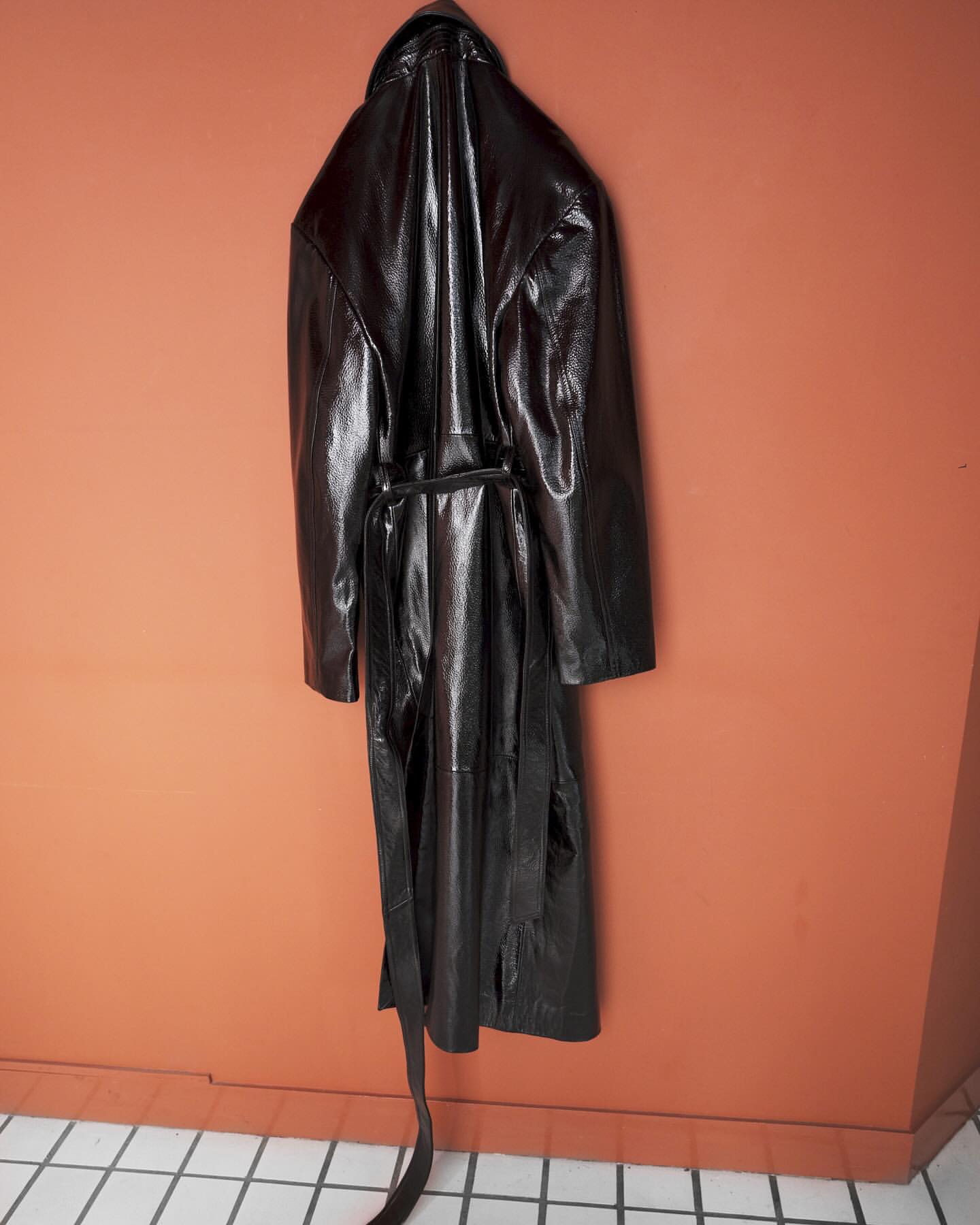 Shiny black long-sleeved coat hanging on a hook in front of an orange wall, with a white tiled floor. Fashionable outerwear.