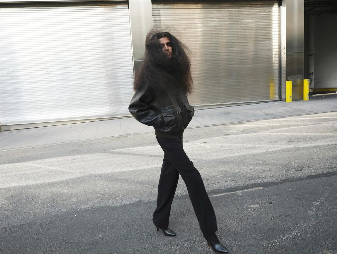 Woman with long dark hair and a leather jacket walks on a city street with a metallic building facade in the background.