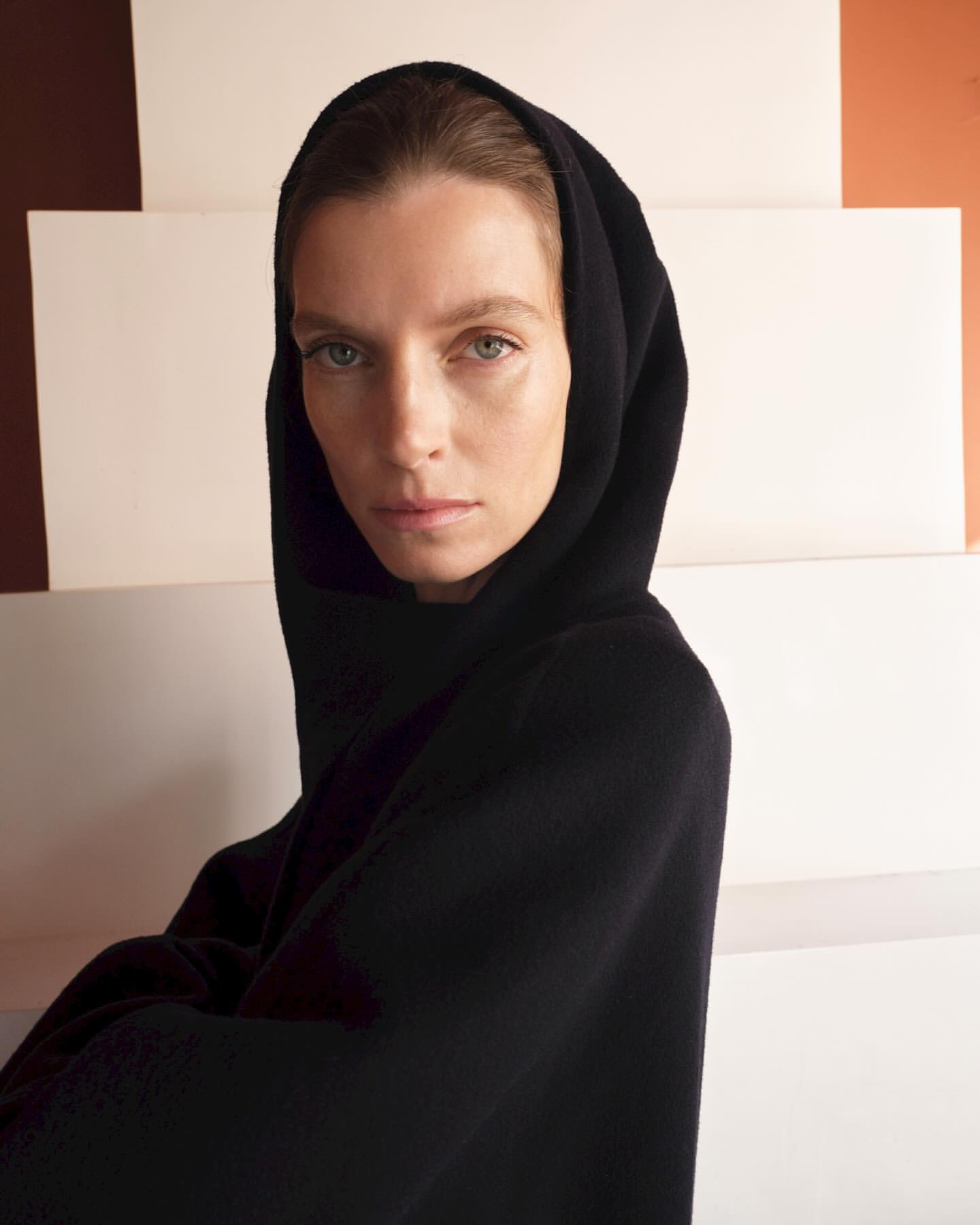 Woman in black hooded garment poses in a studio with geometric backdrop. Close-up portrait captures minimalist fashion.