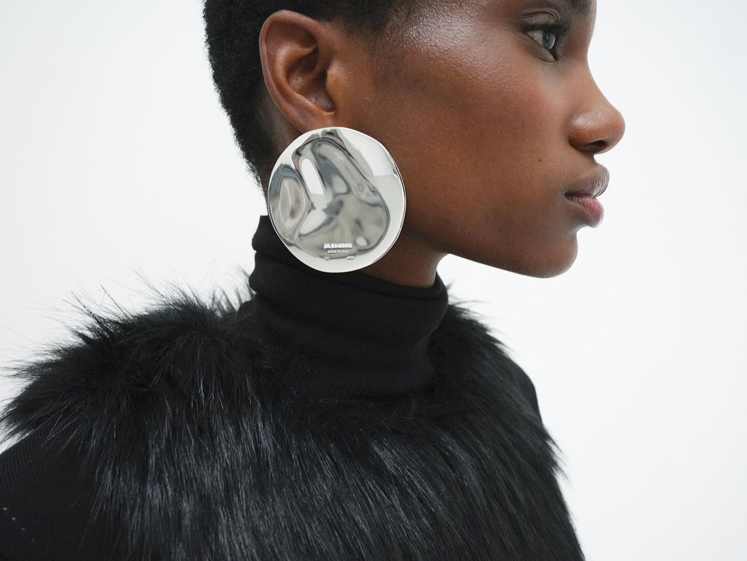 Close-up of an African American fashion model in a fur-trimmed black top, wearing a reflective silver disc earring with logo against white.