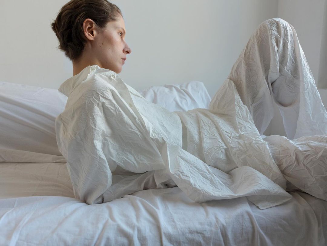 Woman reclines on a white bed covered in textured white sheets in a minimalist indoor setting, looking off to the side.