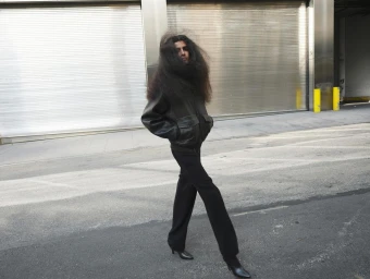 Long-haired person wearing a dark leather bomber jacket and black trousers walking past a corrugated metal rolling shutter in Paris.