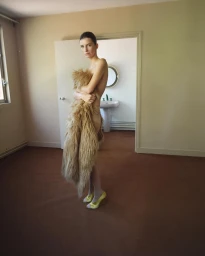 Model wrapped in a shaggy fur stole standing in a room with brown carpet and a doorway revealing a bathroom