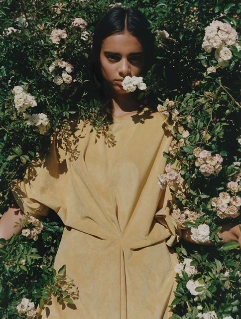 Young woman in a yellow dress surrounded by white roses, posing for a close-up portrait in a natural garden setting with bright lighting.