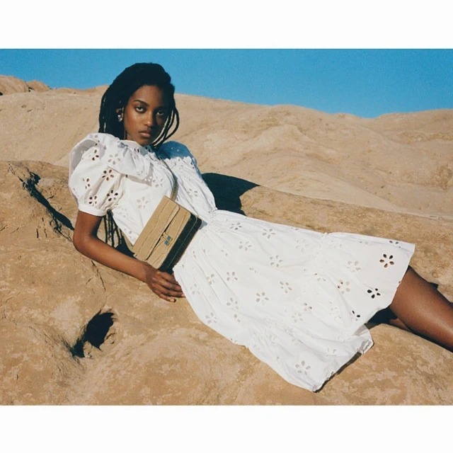 A Black woman in a white dress and shoulder bag poses in a desert landscape with a blue sky. Fashion editorial image.