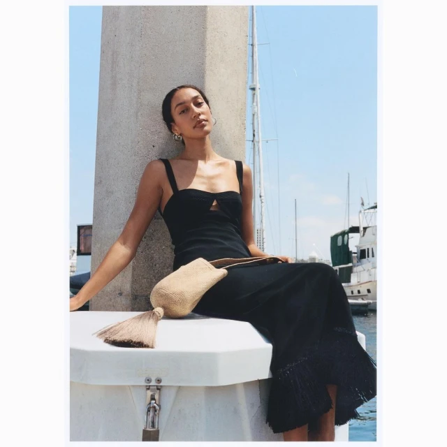 Elegant woman in black dress poses with neutral-tone tassel bag on a boat, with blue sky and water background on a bright day.