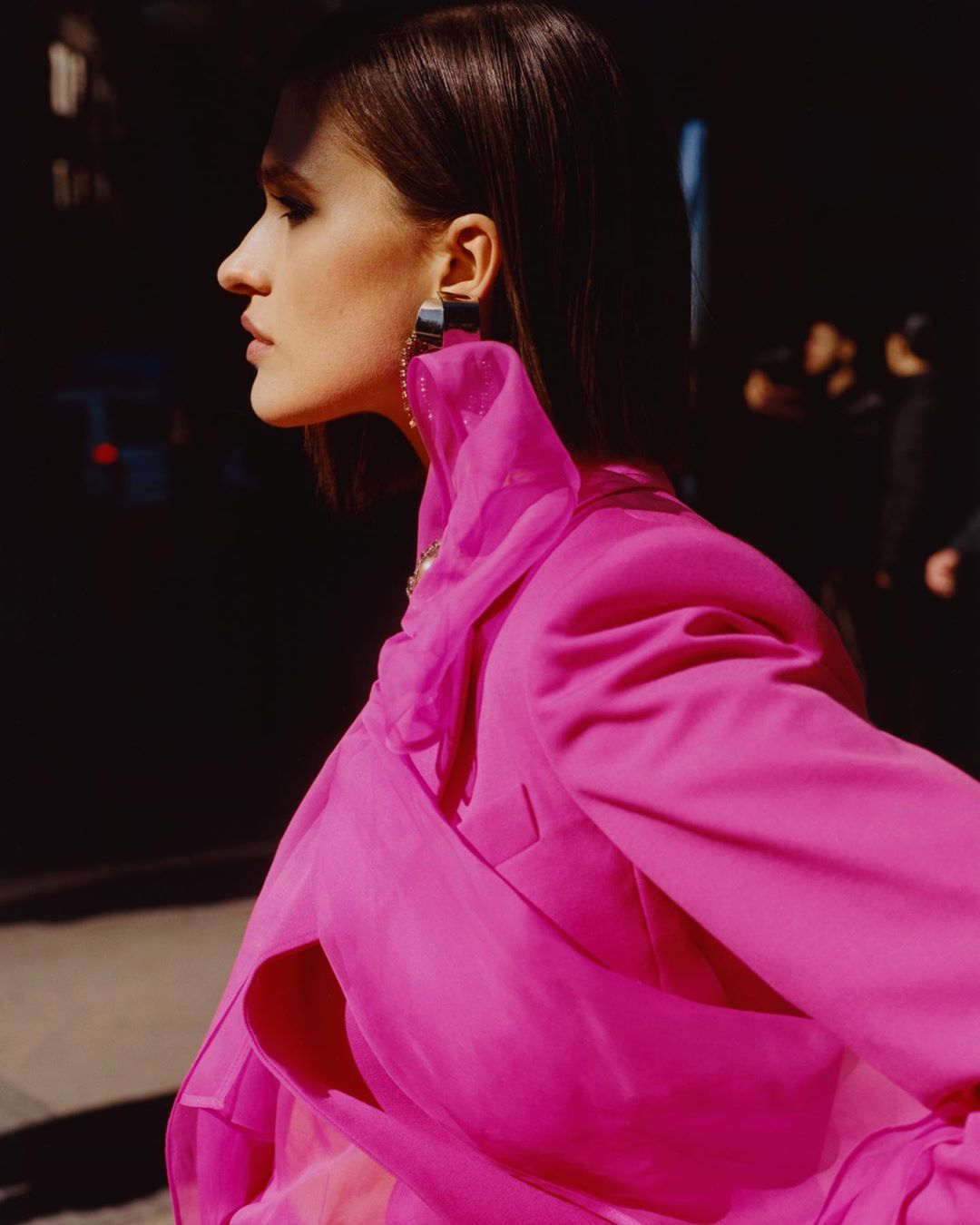Profile shot of a woman in a pink blazer and statement earrings standing outside at night with blurred figures in the background.