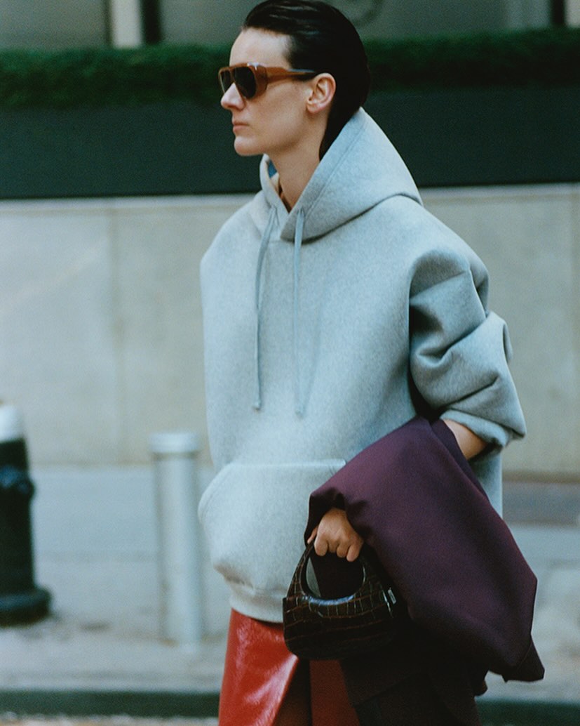 A woman with short dark hair wears sunglasses, a grey hoodie, and a red skirt while holding a small dark handbag on a city street.