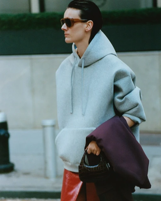 A woman with short dark hair wears sunglasses, a grey hoodie, and a red skirt while holding a small dark handbag on a city street.