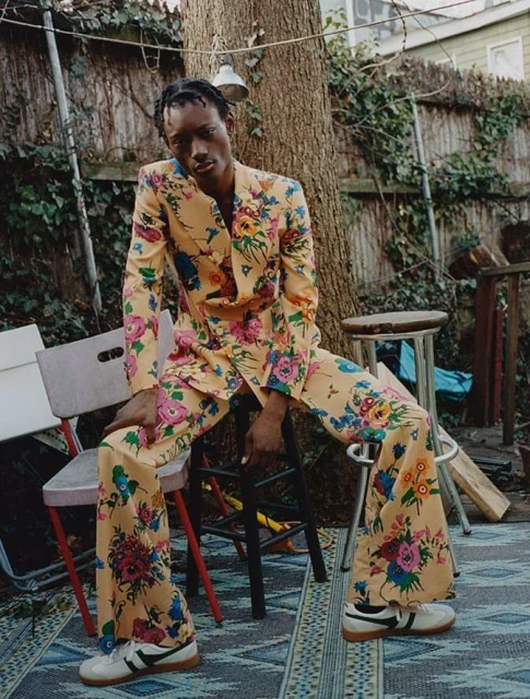 Man poses in a floral suit outdoors, sitting on a stool with chairs and a patterned rug, near a tree and wooden fence.