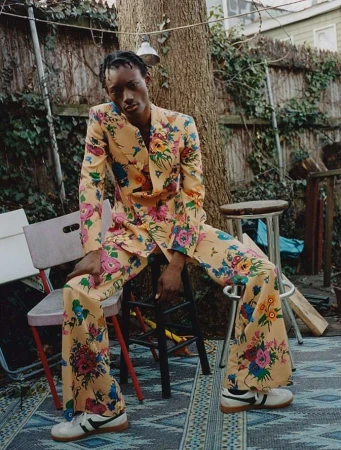Man poses in a floral suit outdoors, sitting on a stool with chairs and a patterned rug, near a tree and wooden fence.
