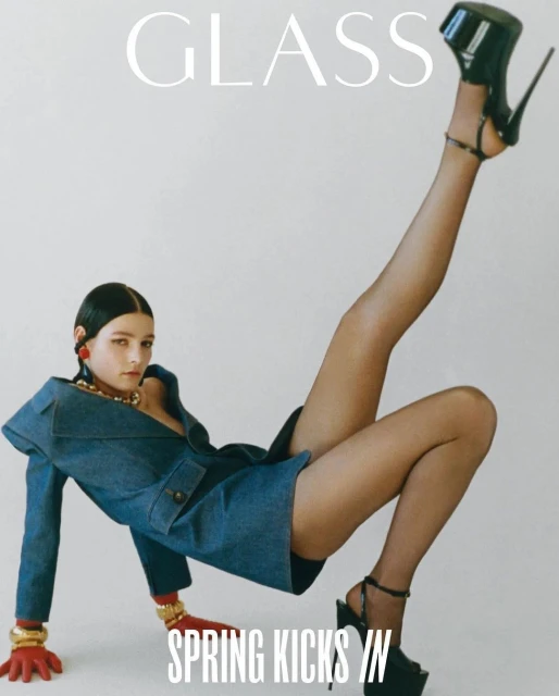 Editorial fashion shot of a woman in a denim suit jacket, platform heels, posing on the floor in a photo studio with white background.