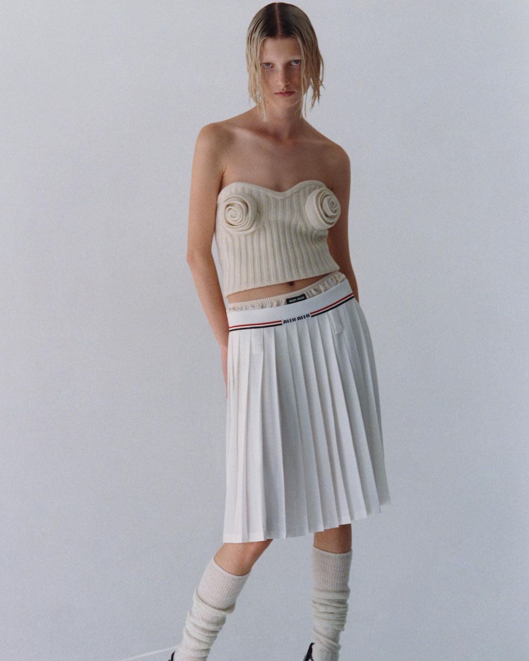 A female model poses in a studio wearing a knit top with floral details, a pleated skirt, and tube socks against a white backdrop.