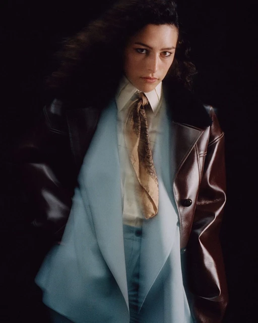 Adult female with curly hair poses wearing a brown leather jacket, light blue suit, off-white shirt, and patterned tan tie against dark backdrop.