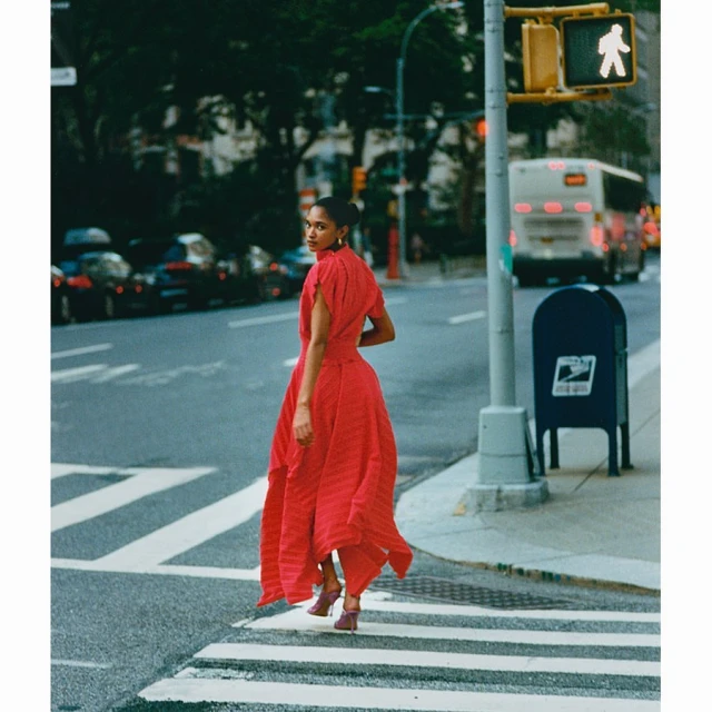 A woman in a red dress crosses a city street at a pedestrian crossing, captured in a medium shot with a vintage fashion aesthetic.