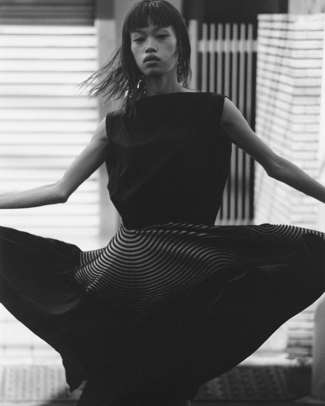 Monochrome fashion shot of an Asian woman in a sleeveless top and striped skirt, standing outdoors with her arms outstretched.