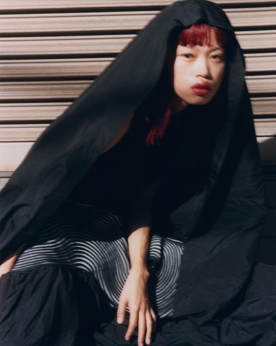 Fashion portrait of a red-haired person with dark makeup, under a black hood and patterned dress, seated against a metal wall.