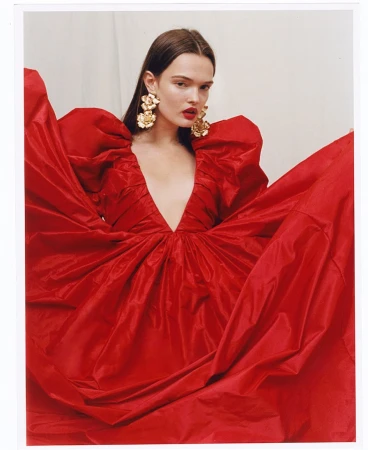 A woman wearing a vibrant red dress with voluminous sleeves and large gold floral statement earrings against a neutral backdrop
