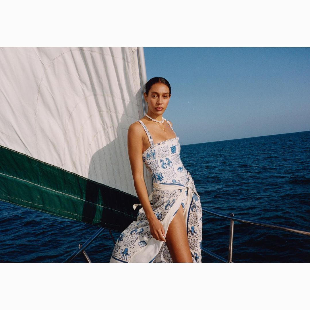 Woman wearing a printed swimsuit and wrap stands on a sailboat with blue water and sky in the background on a sunny day.