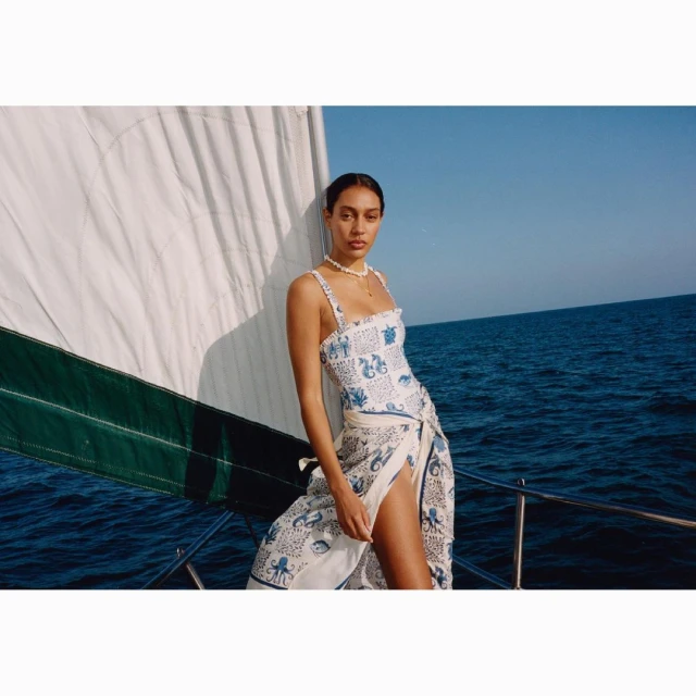 Woman wearing a printed swimsuit and wrap stands on a sailboat with blue water and sky in the background on a sunny day.