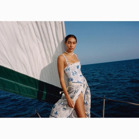Woman wearing a printed swimsuit and wrap stands on a sailboat with blue water and sky in the background on a sunny day.