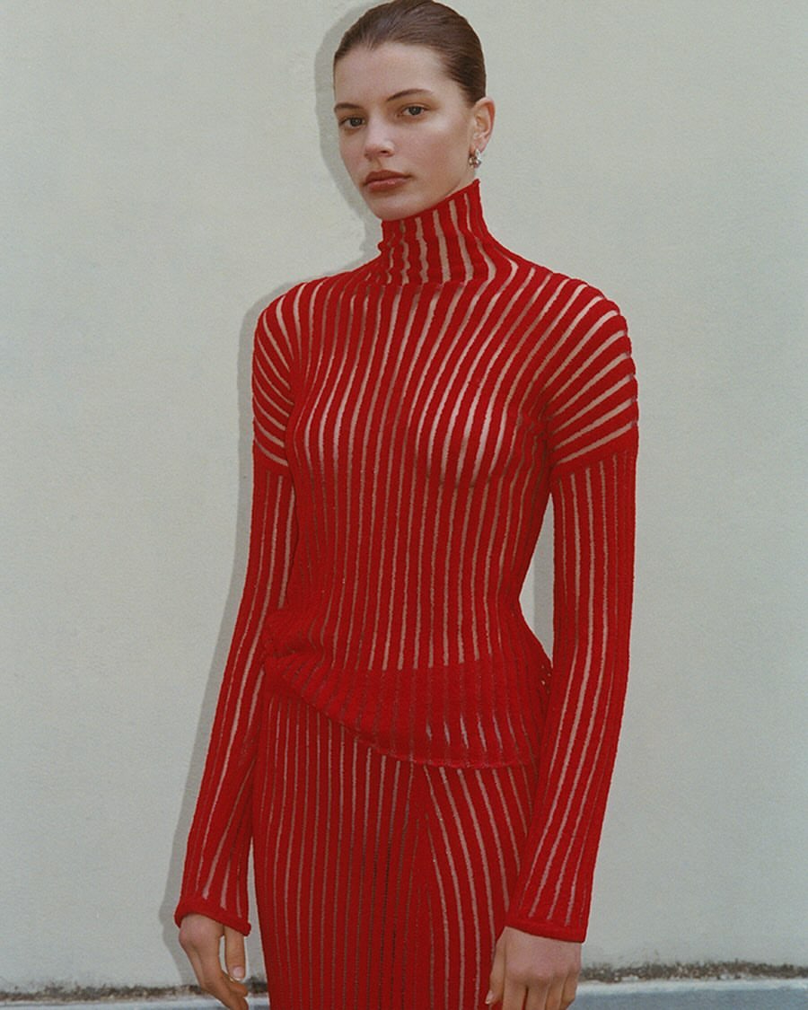 An adult female model poses indoors wearing a semi-sheer vertical striped red turtleneck and matching red skirt against a neutral wall.