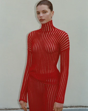 An adult female model poses indoors wearing a semi-sheer vertical striped red turtleneck and matching red skirt against a neutral wall.
