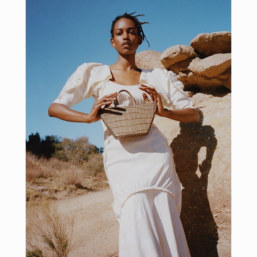 A woman in a white dress stands outdoors holding a brown faux-crocodile leather purse against a backdrop of rocks and blue sky.