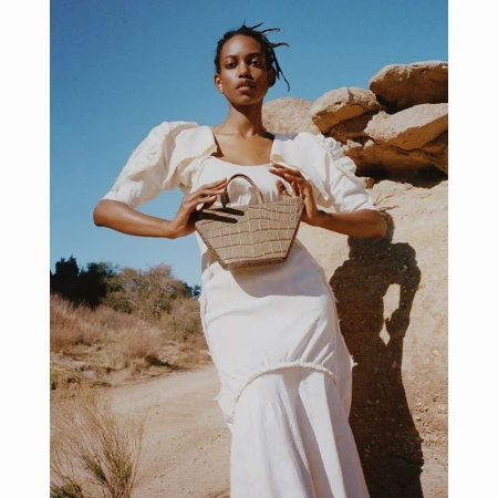 A woman in a white dress stands outdoors holding a brown faux-crocodile leather purse against a backdrop of rocks and blue sky.