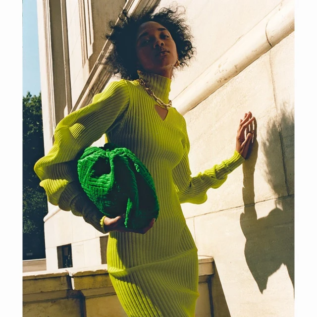 Woman in lime green dress holds green handbag against sunlit wall, accessorized with gold chain. Fashion shot with natural light.