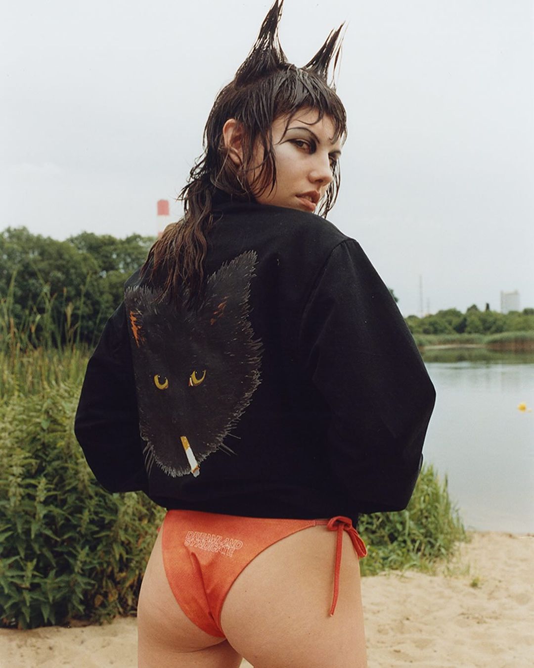 Young woman with unique hairstyle and black jacket featuring a cat graphic, posing outdoors near a lake on a cloudy day.