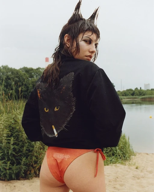 Young woman with unique hairstyle and black jacket featuring a cat graphic, posing outdoors near a lake on a cloudy day.