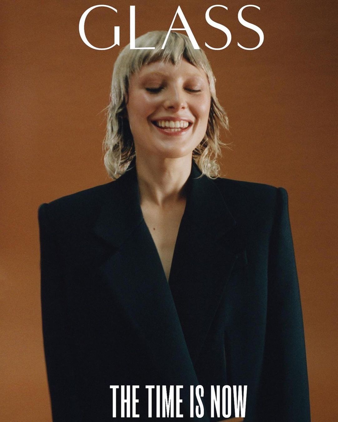 Smiling woman with blonde fringe wearing a black blazer on a solid tan background. 'The Time Is Now' text overlays the bottom.