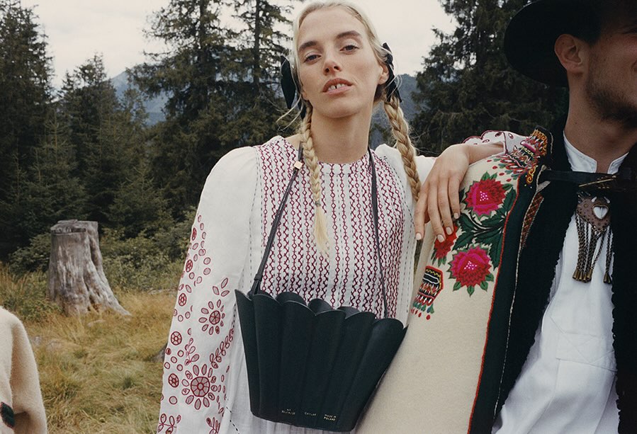 A woman with blonde braids and a black purse poses next to a man wearing traditional clothing against a forest and mountain backdrop.