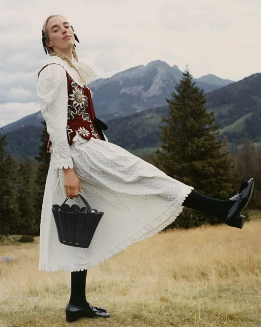 Woman in traditional dress with bucket bag strikes a pose outdoors. Mountain landscape and cloudy sky in the background. 