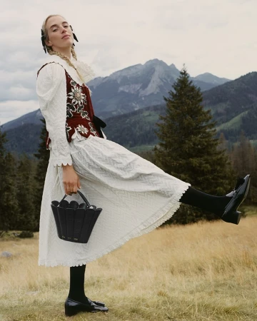 Woman in traditional dress with bucket bag strikes a pose outdoors. Mountain landscape and cloudy sky in the background. 