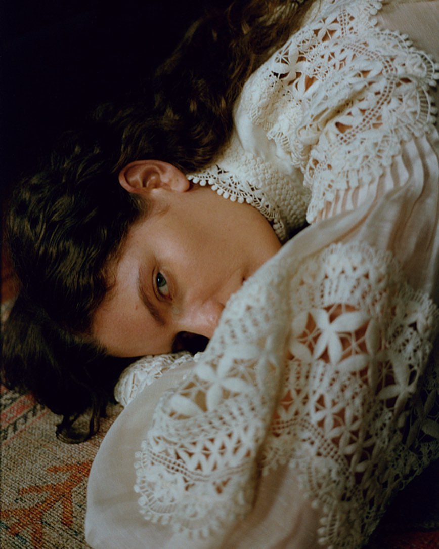 A young woman with curly hair rests her head on a patterned surface, showcasing a delicate white lace dress in a softly lit scene.