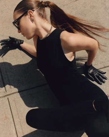 Model in black sleeveless top and leather gloves with a long ponytail posing against a concrete ground outdoors