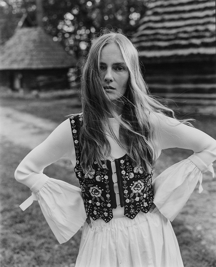 Portrait of a blonde woman in a folk-style embroidered vest, wearing a long-sleeve shirt, posing outdoors with a blurred landscape background.