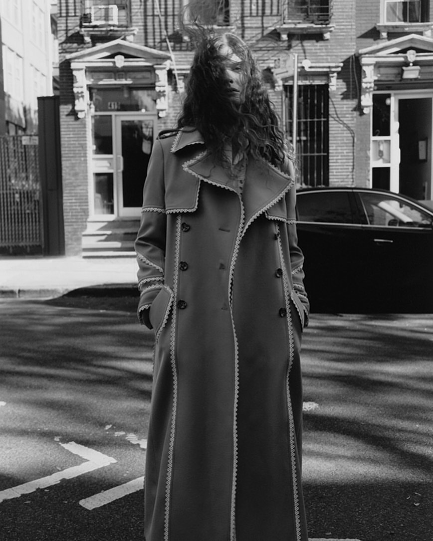 A black and white fashion portrait of a woman in a long coat standing on a city street, with buildings and a car in the background.