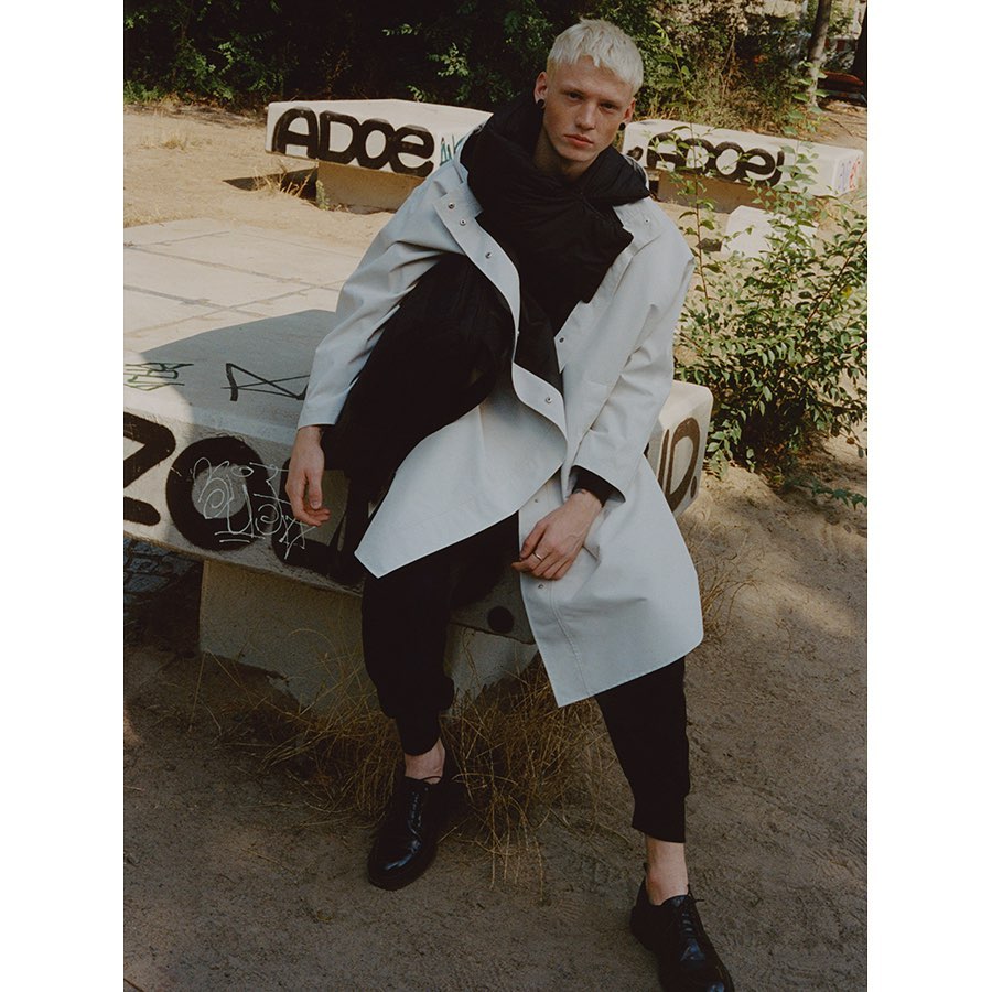 Man in off-white trench coat and black scarf sits on concrete bench outdoors with graffiti, showcasing fashion in urban setting.