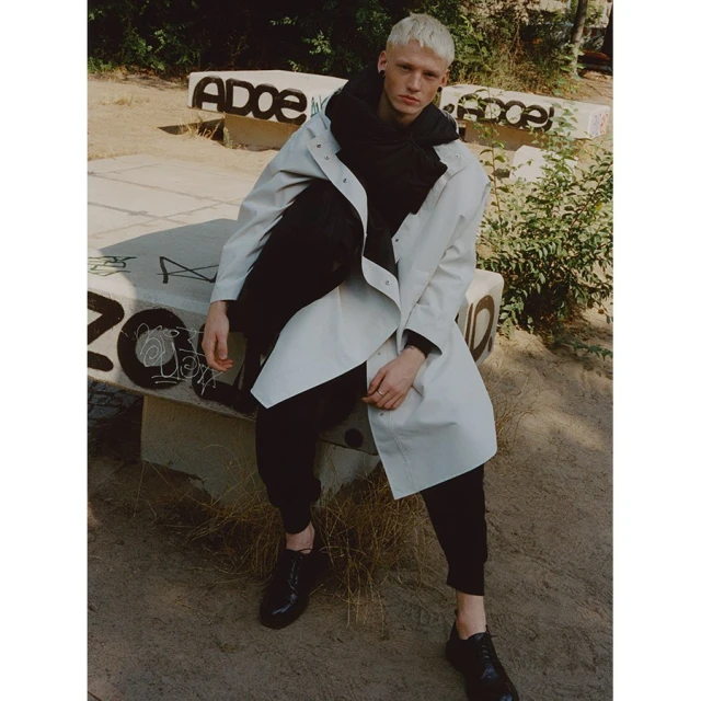 Man in off-white trench coat and black scarf sits on concrete bench outdoors with graffiti, showcasing fashion in urban setting.