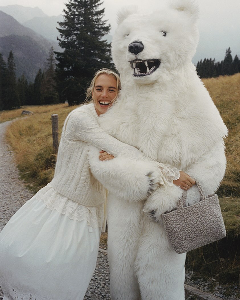 Woman smiles, hugging a polar bear mascot in a mountainous setting. She wears a white dress; the mascot has realistic bear features.