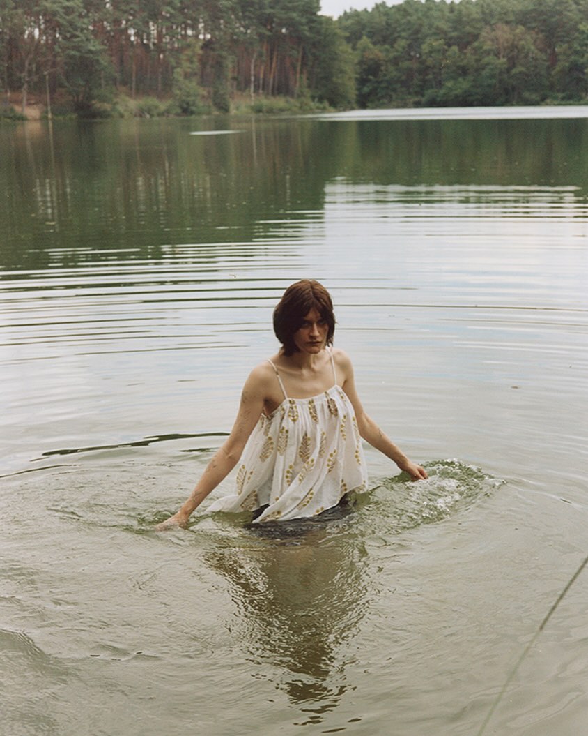 Woman stands in lake water with a forest backdrop. She wears a light-colored floral print top, creating a serene outdoor scene.