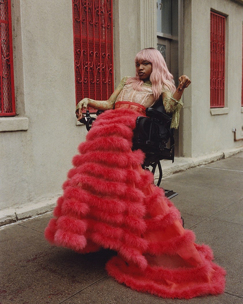 Adult female model with pink hair poses in a wheelchair wearing a tiered pink dress in front of a white building with red window frames.