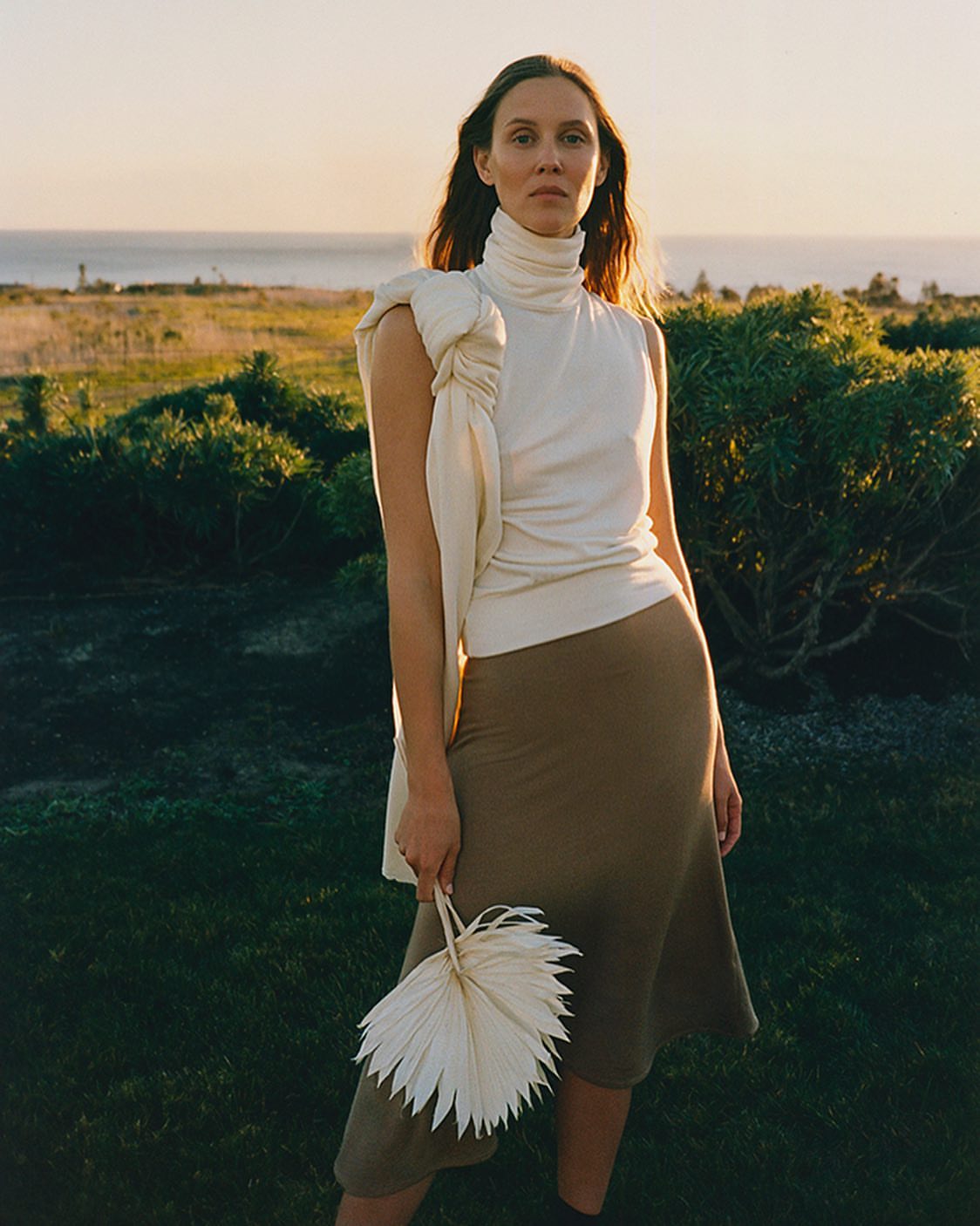 A woman in a white top and skirt poses outdoors in a grassy area, holding a palm fan, with the ocean and sky visible in the background.