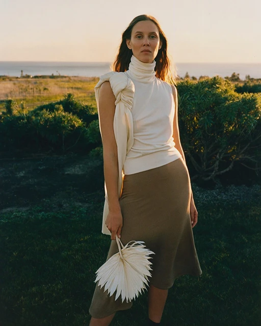 A woman in a white top and skirt poses outdoors in a grassy area, holding a palm fan, with the ocean and sky visible in the background.