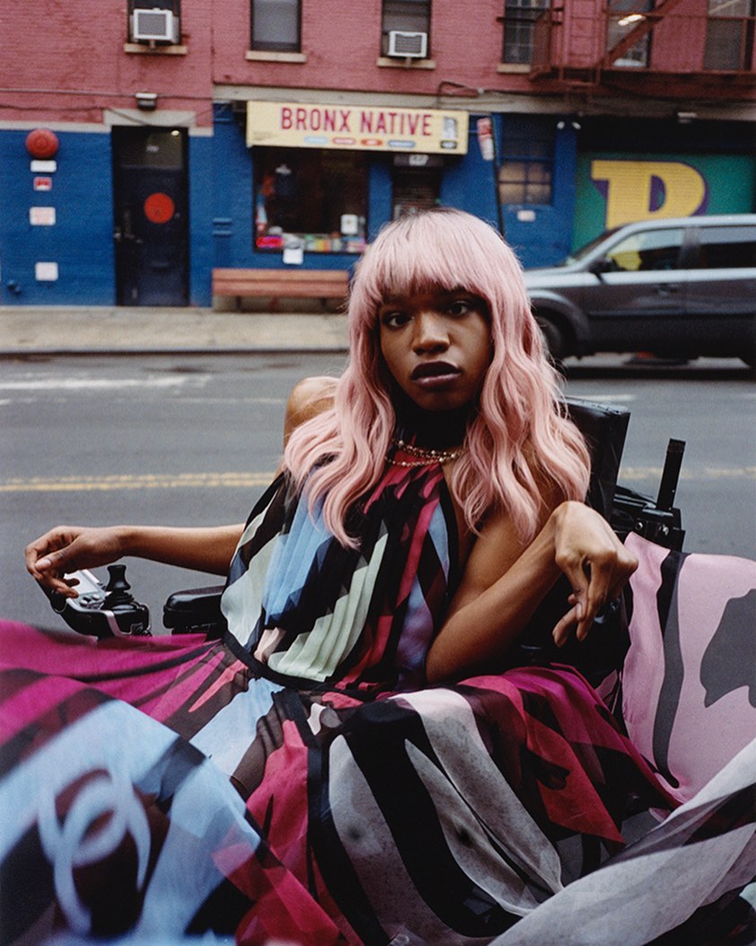 A stylish woman in a wheelchair sits in a city street with a blue and red building in the background, bathed in natural light.
